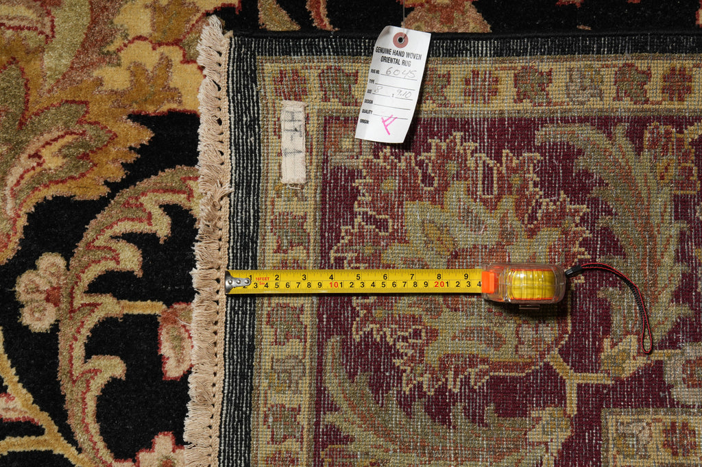 A vintage patterned area rug with a dark charcoal border and fringed edge, shown with a yellow tape measure laid out horizontally to indicate its dimensions.