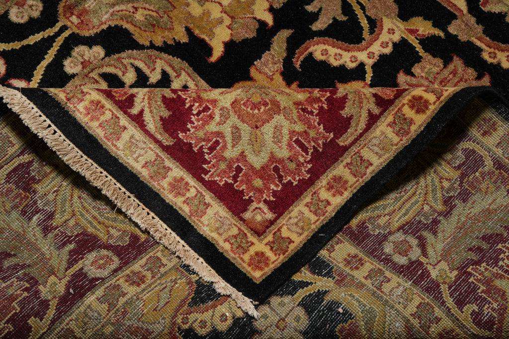 Close-up of a patterned area rug corner showing a deep red central field, beige and gold floral motifs, black borders, and cream fringe, layered over other rugs.