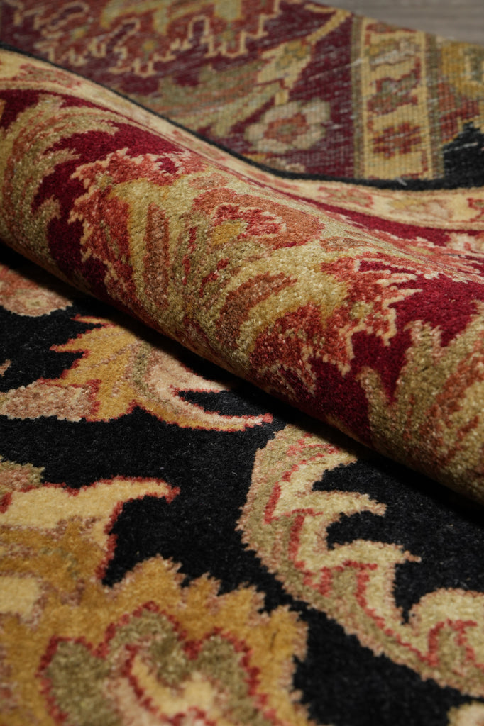 A close-up of a folded area rug showing its detailed pattern and pile texture. The rug features a complex traditional design with a black base, prominent gold and beige floral motifs, and deep red sections.