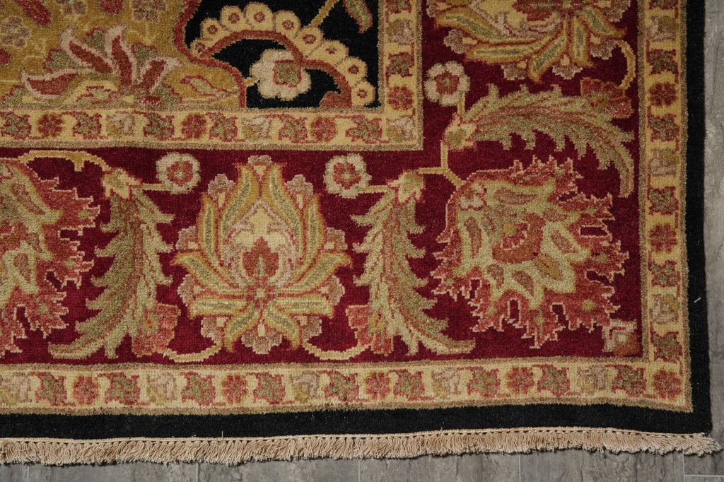 Close-up of an ornate patterned area rug corner showing deep red, beige, black, and muted green floral motifs with fringed edge and woven texture.