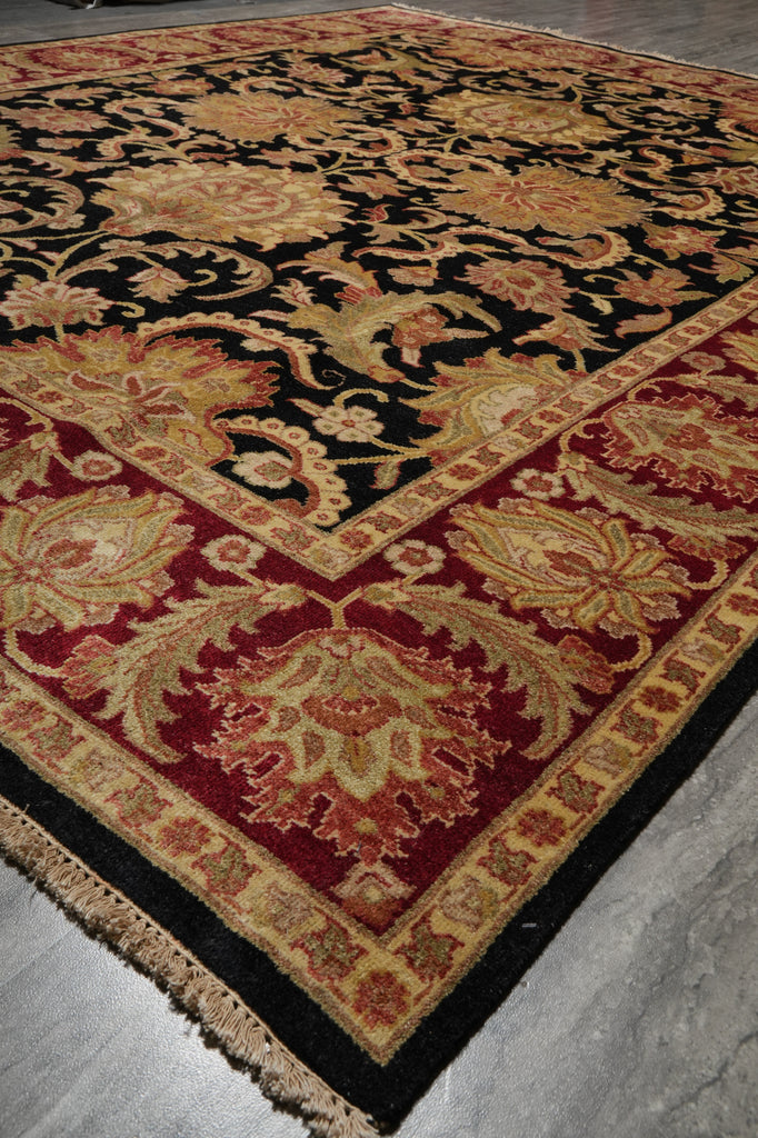 A traditional oriental-style area rug with an intricate floral and scroll pattern, featuring a black field, deep red borders, and beige and gold accents, laid out on a gray concrete floor.