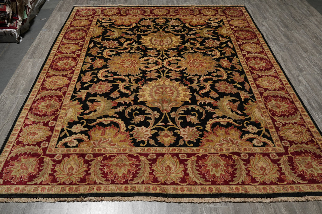 A large rectangular area rug with an ornate traditional floral pattern, featuring a black center field, deep red borders, and gold and beige details, laid out on a gray wood floor.