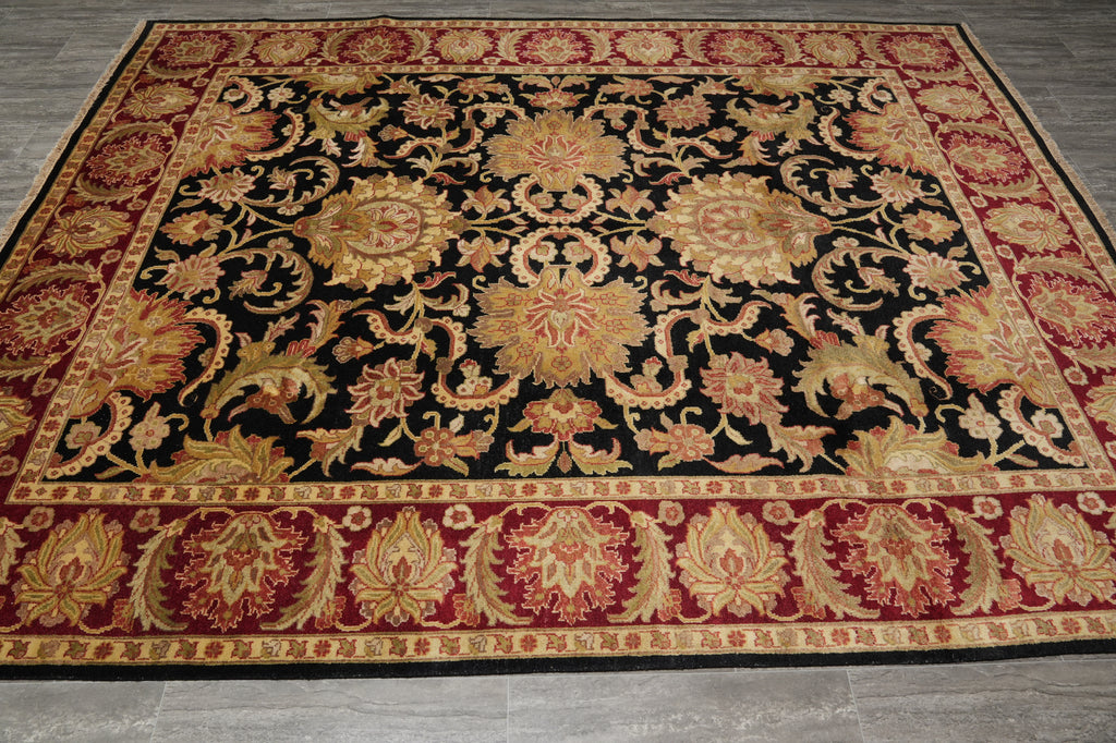 An ornate rectangular area rug with a black center field, featuring large floral medallions and scrolling vine motifs in gold, red, and beige, surrounded by a detailed patterned border, shown on a gray tiled floor.