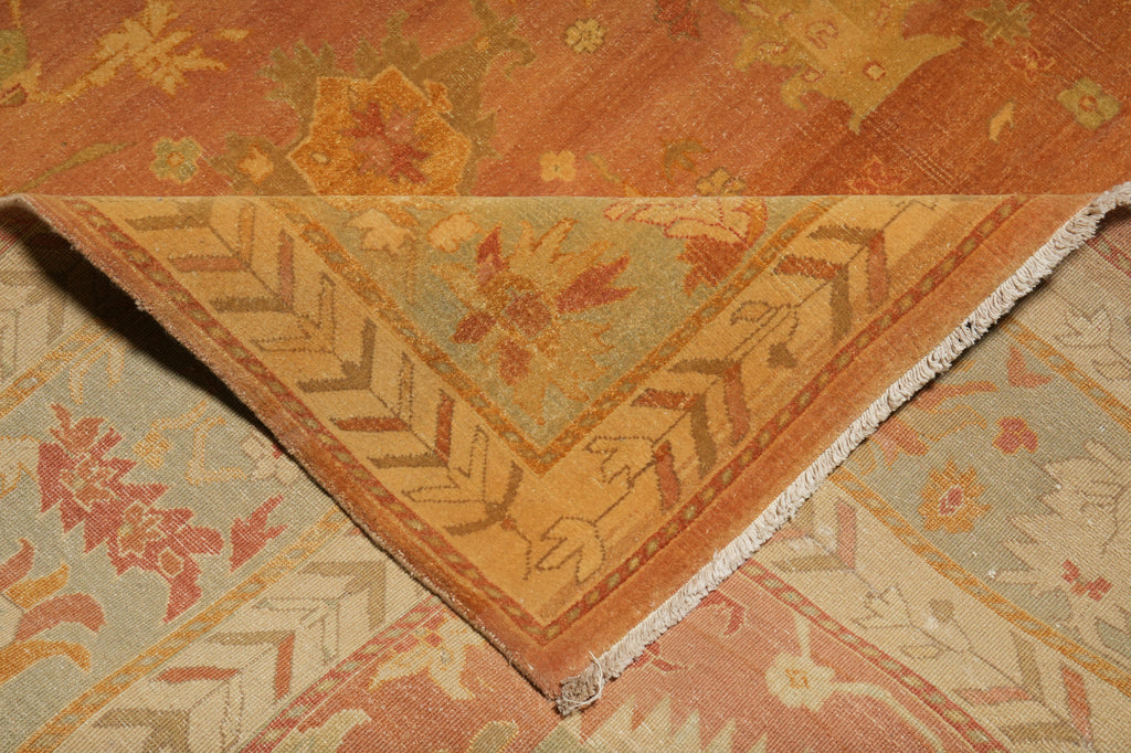 A close-up of a folded area rug showing its corner and fringe, featuring a warm orange and beige floral pattern over a muted sage green and cream geometric border.