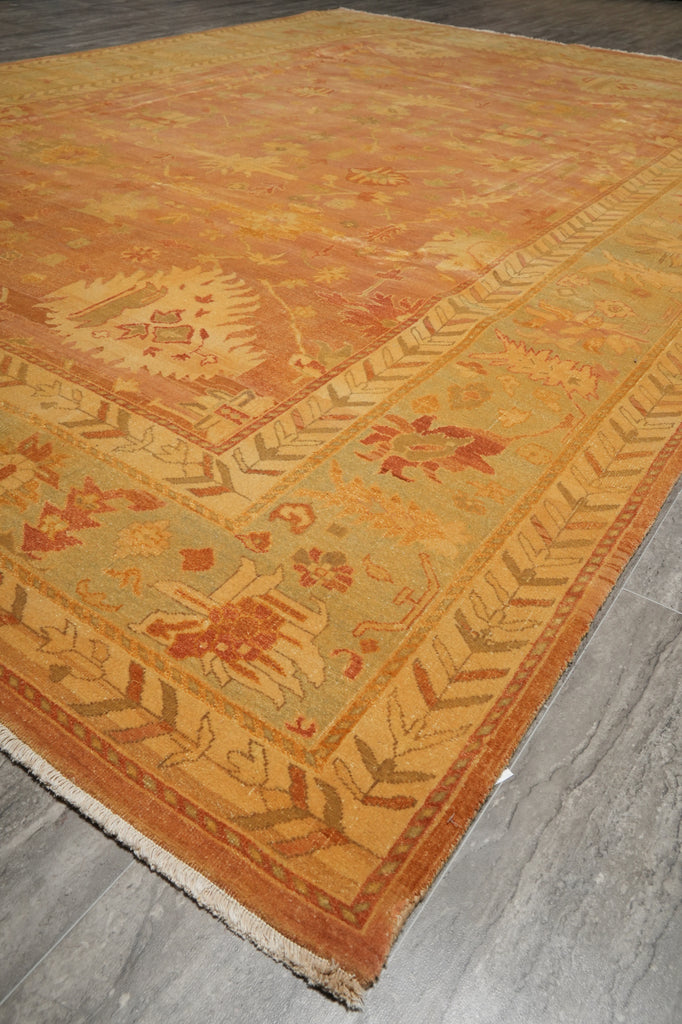 A square area rug with a traditional floral pattern in warm shades of ochre, mustard, and terracotta, laid out on a gray wood floor.