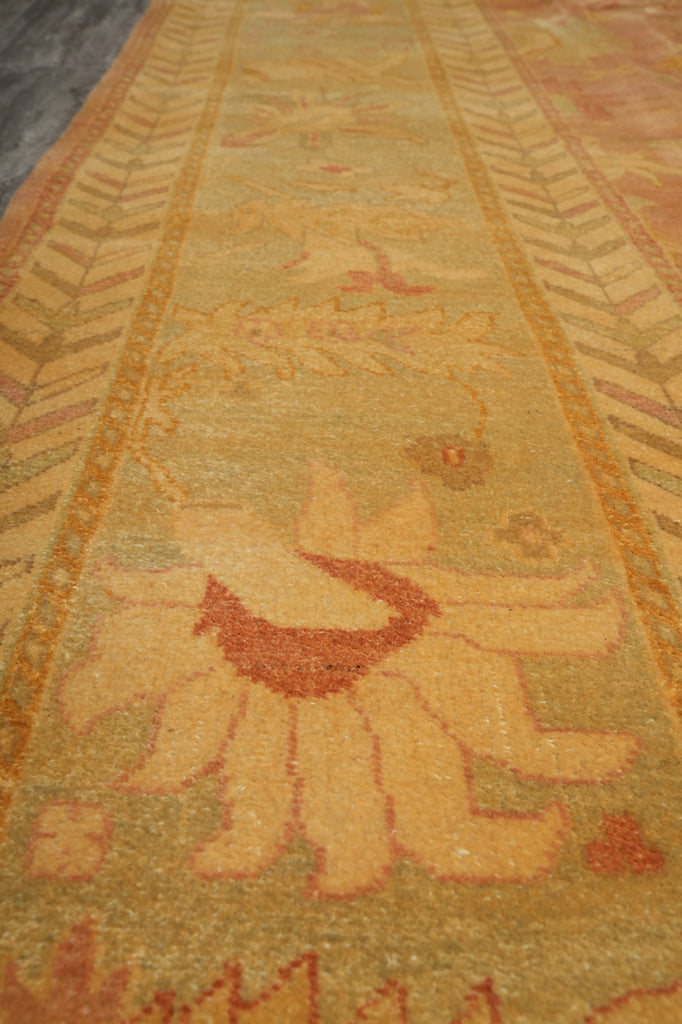 A close-up view of a patterned area rug with a warm mustard yellow and beige base, featuring a central floral motif in rust red and orange, bordered by geometric patterns.