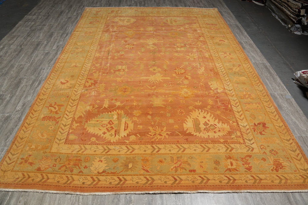 A large rectangular area rug with an intricate traditional pattern, featuring a warm orange and golden-yellow center with a beige border, laid out on a gray wood floor.
