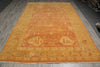 Load image into Gallery viewer, A large rectangular area rug with an intricate traditional pattern, featuring a warm orange and golden-yellow center with a beige border, laid out on a gray wood floor.