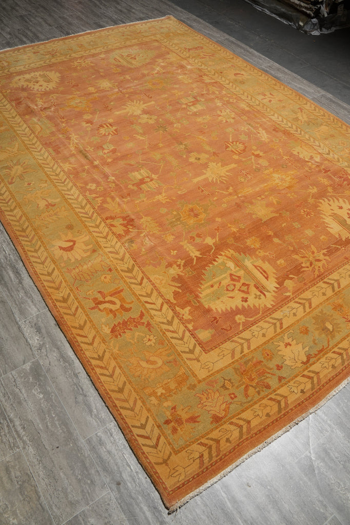 A rectangular area rug with a warm orange and beige central field and a contrasting olive green border, laid out on a gray wood floor.