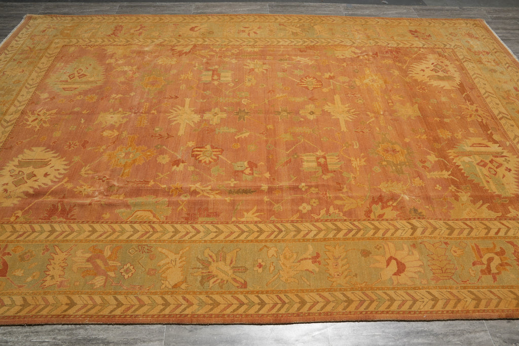 An antique-style oriental area rug with a faded orange and ochre center field, decorated with traditional motifs and a detailed patterned border, laid out on a dark gray tiled floor.