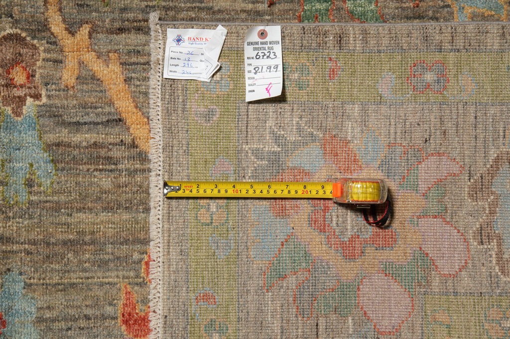 A close-up of a patterned area rug with a measuring tape laid across it and two inventory tags attached near the top edge, showing the rug's dimensions.
