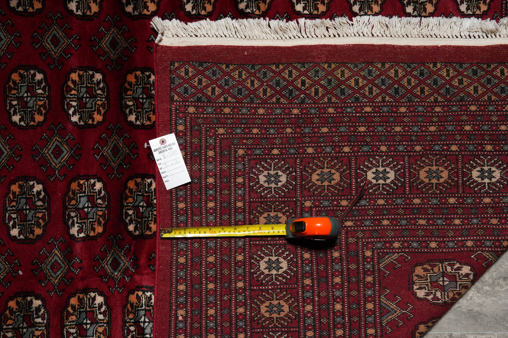 A traditional patterned area rug in deep red with geometric and floral motifs, shown with a yellow tape measure and a white product tag on a dark surface.