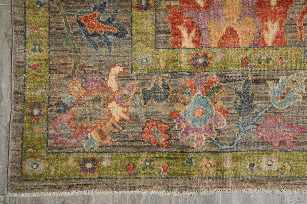 A close-up of a traditional patterned rug corner showing a muted olive green border, a gray-brown field, and floral motifs in rust red, dusty blue, lavender, and peach.