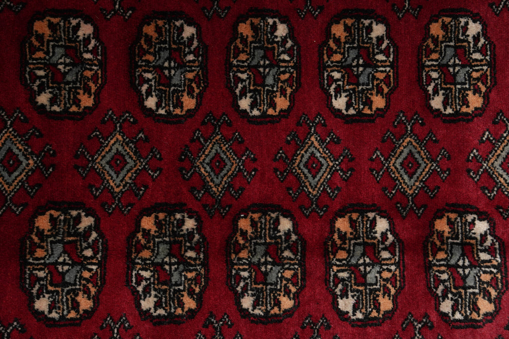 A close-up of a red patterned rug featuring repeating geometric medallion motifs in beige, black, blue, and red.