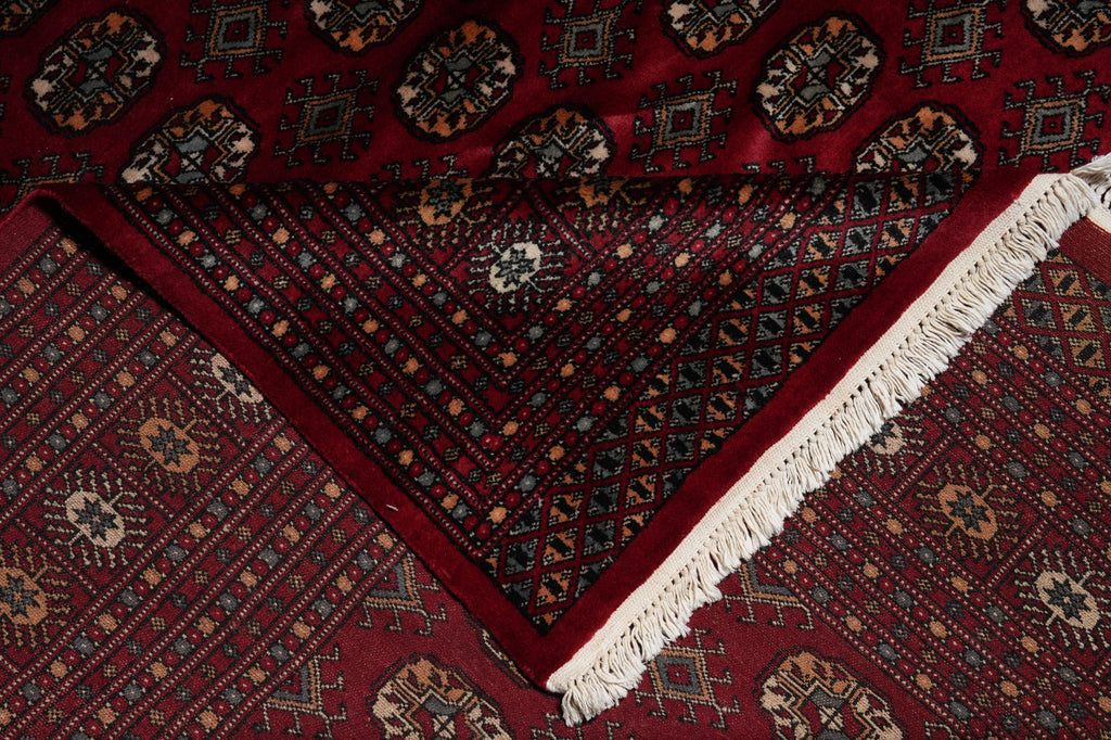 A close-up of a traditional patterned rug with a deep red background, intricate geometric and floral motifs in black, beige, and blue, and a white fringed edge.