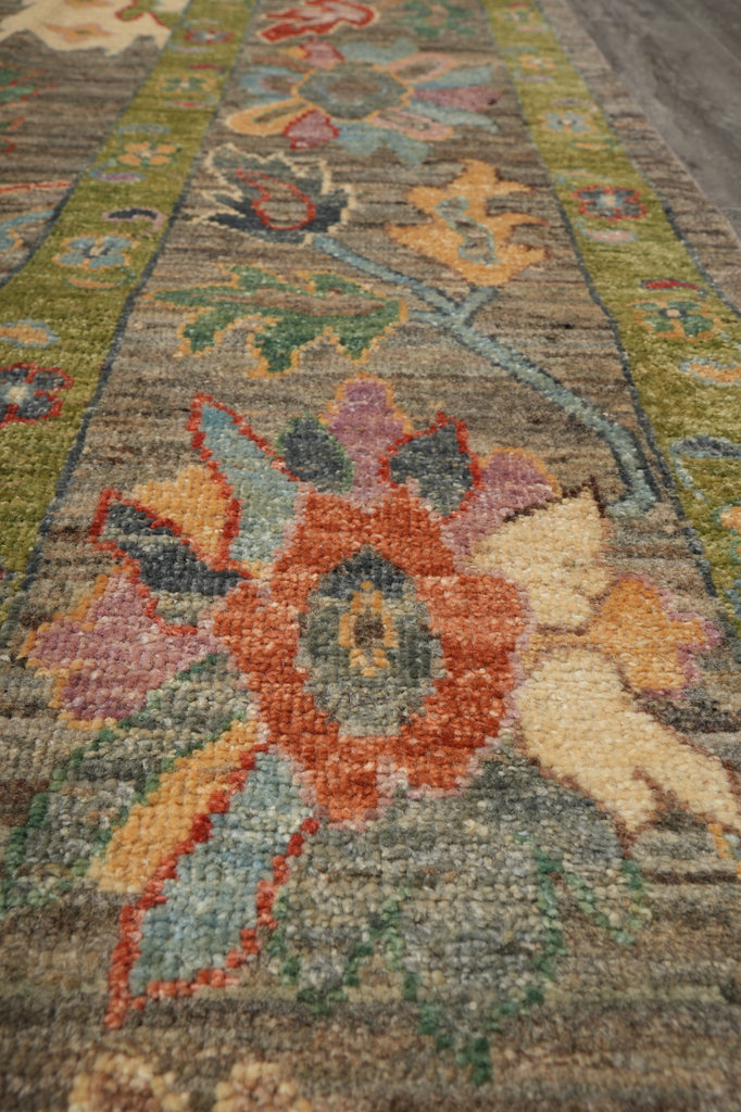 A close-up view of a traditional area rug with a floral pattern in muted earthy tones, featuring a central rust-orange flower, beige cherub motifs, and green borders.