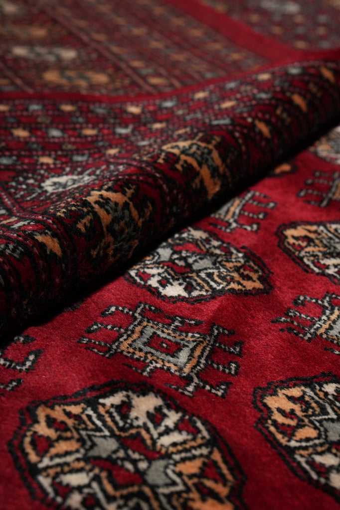 A close-up of a folded patterned rug or textile with a deep red field and ornate geometric motifs in beige, black, and muted blue.