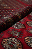 Load image into Gallery viewer, A close-up of a folded patterned rug or textile with a deep red field and ornate geometric motifs in beige, black, and muted blue.