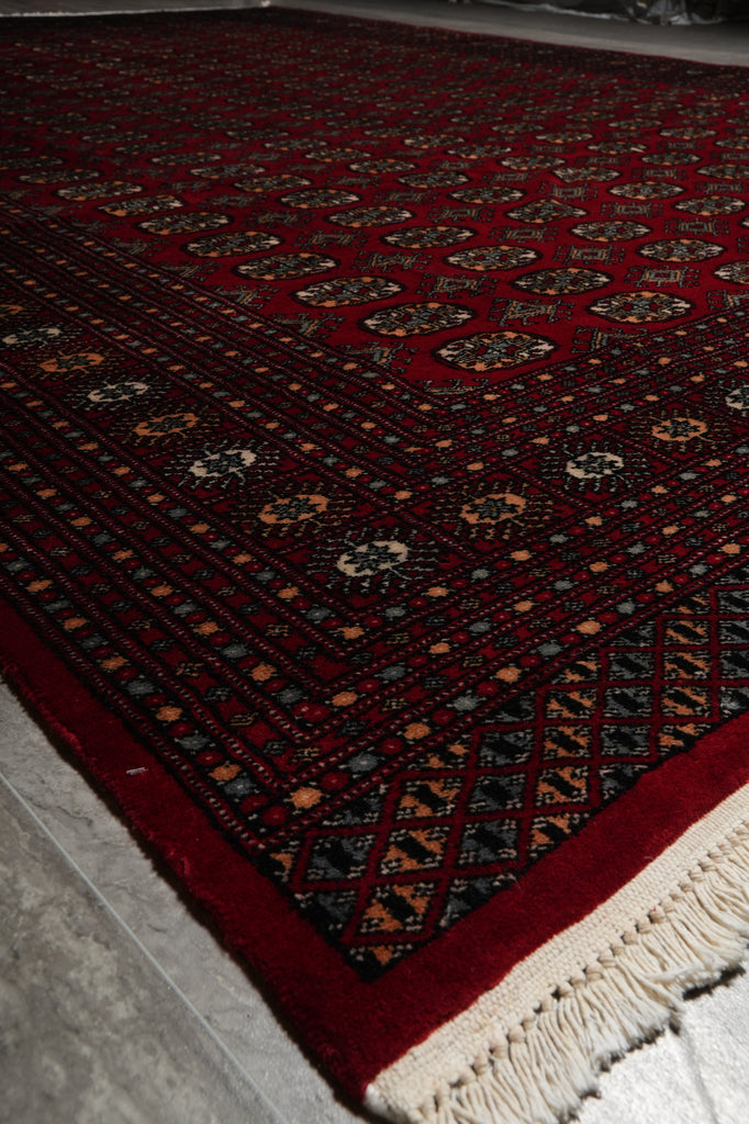 A close-up of a traditional patterned rug with a deep red field, cream fringe, and dark navy and beige geometric motifs.