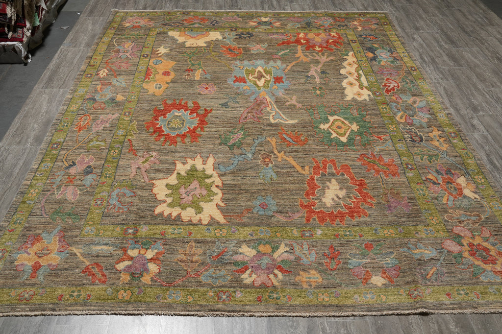 A rectangular area rug with a traditional floral pattern, displayed on a gray wood floor. The rug features a muted olive-green and brown base with prominent red, beige, and light blue floral motifs.