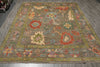 Load image into Gallery viewer, A rectangular area rug with a traditional floral pattern, displayed on a gray wood floor. The rug features a muted olive-green and brown base with prominent red, beige, and light blue floral motifs.
