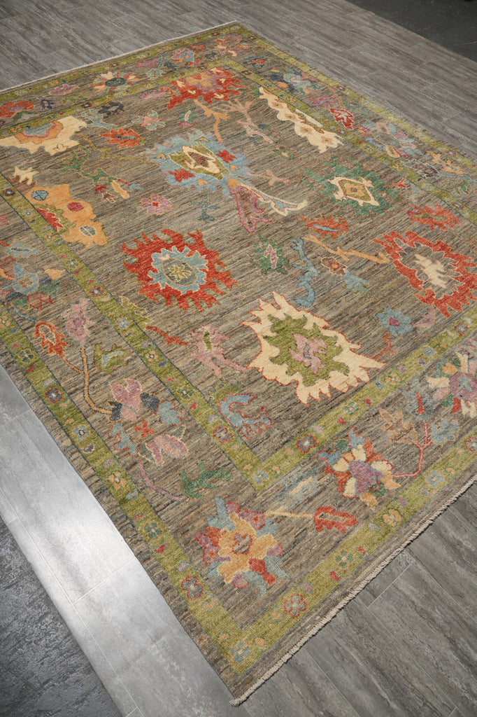 A rectangular area rug with an intricate vintage-style floral and medallion pattern in muted earthy tones, shown laid out on a gray wood-plank floor.