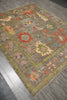 Load image into Gallery viewer, A rectangular area rug with an intricate vintage-style floral and medallion pattern in muted earthy tones, shown laid out on a gray wood-plank floor.