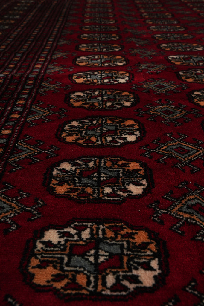 A close-up view of a traditional patterned rug with a deep red background and repeating geometric medallion motifs in beige, black, and muted blue-gray.