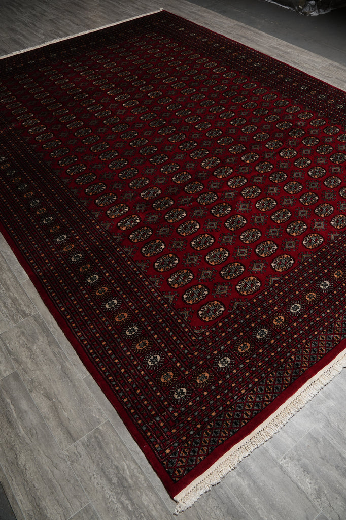 A traditional oriental area rug with a deep red field, intricate geometric patterns, and cream fringe, laid out on a gray wood-look floor.