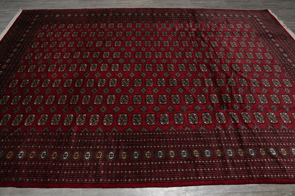 A large traditional Persian-style area rug with a deep red field, featuring rows of small geometric medallions and a detailed patterned border, laid flat on a gray floor.