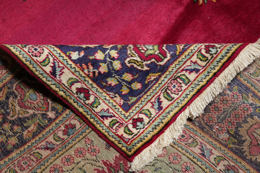 A close-up of a patterned area rug corner showing a deep navy blue central field with floral motifs, surrounded by cream, red, and green borders, with cream fringe along one edge. Another magenta rug is partially visible at the top.