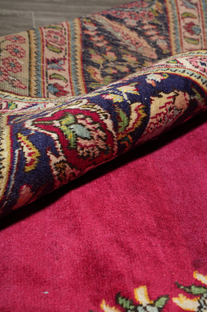 A close-up of a folded area rug showing a rich magenta-pink field with a traditional multicolor pattern in navy, red, green, and cream, laid over a gray wood floor.