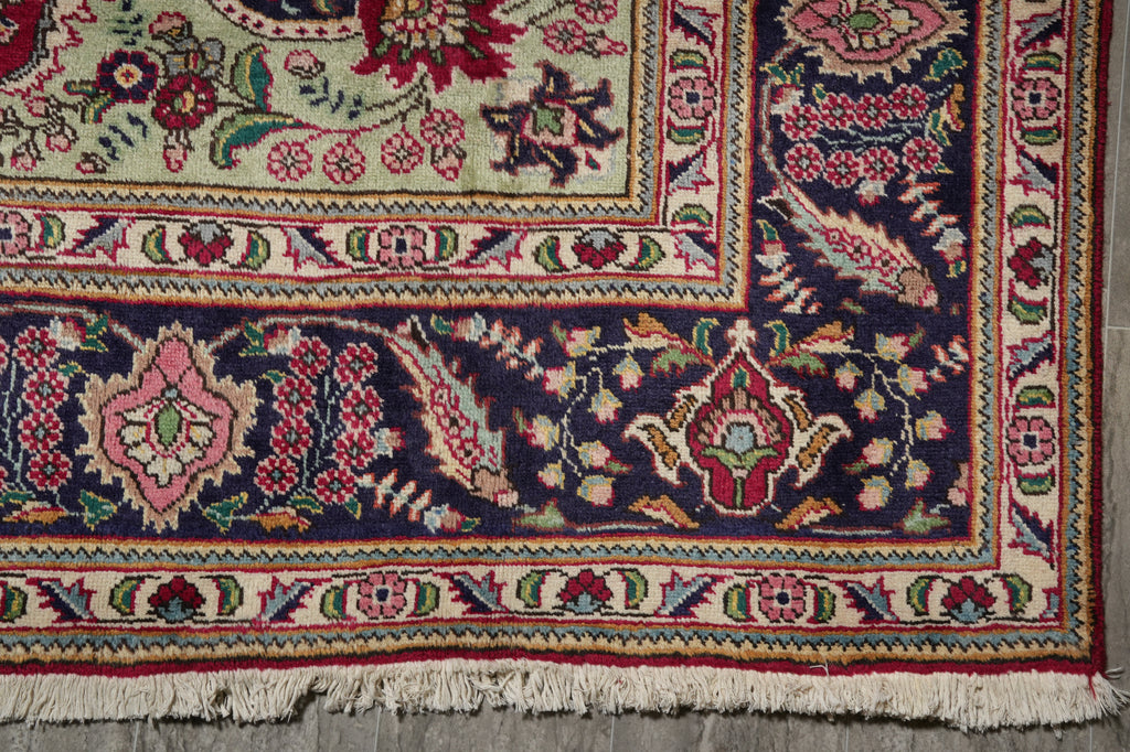 A close-up of an ornate patterned rug corner showing cream fringe, a dark navy field with floral motifs, and borders in pink, red, green, and beige.