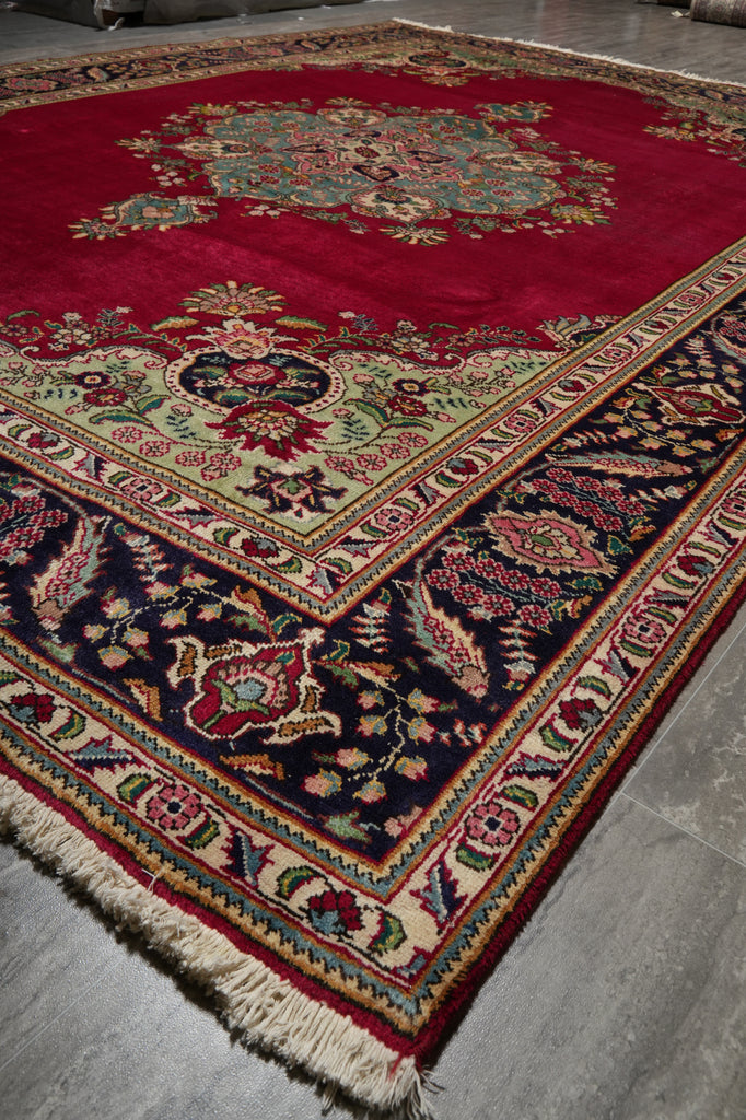 A traditional oriental area rug with a deep red field, ornate floral medallion, and dark navy border, shown laid out on a gray concrete floor.