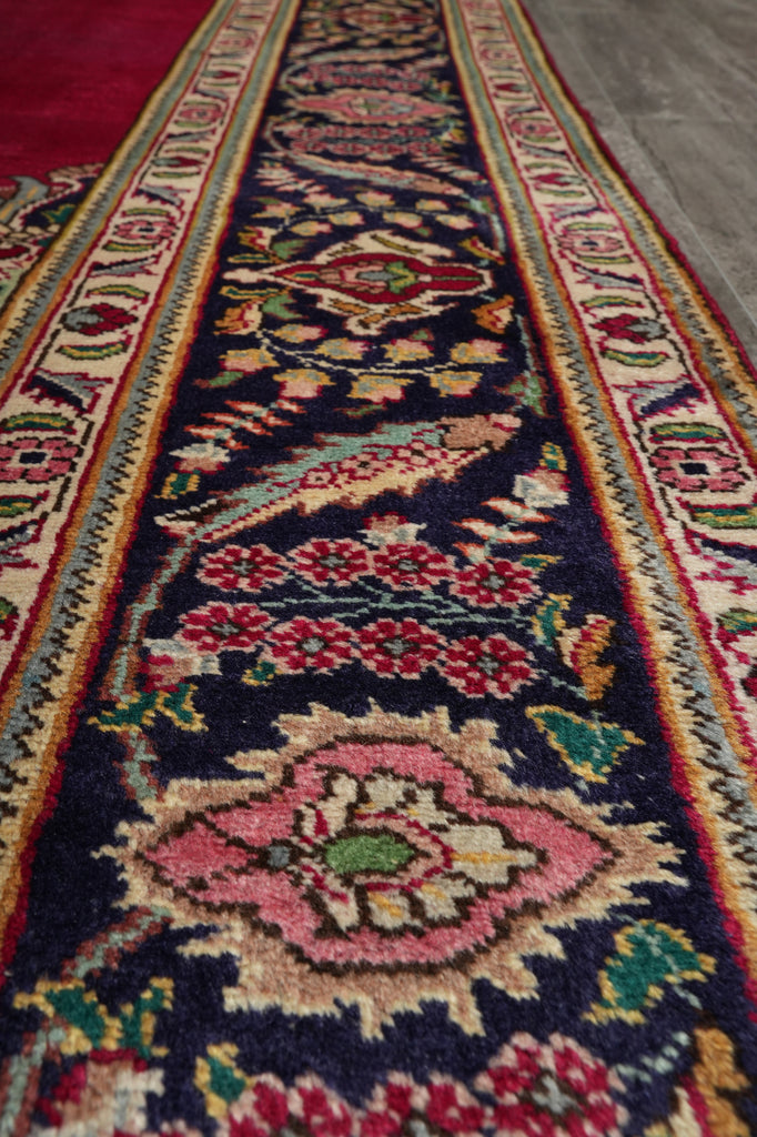 A close-up view of a traditional patterned area rug runner with a dark navy blue center, featuring intricate floral and bird motifs in pink, red, green, and beige, bordered by red and cream patterns.