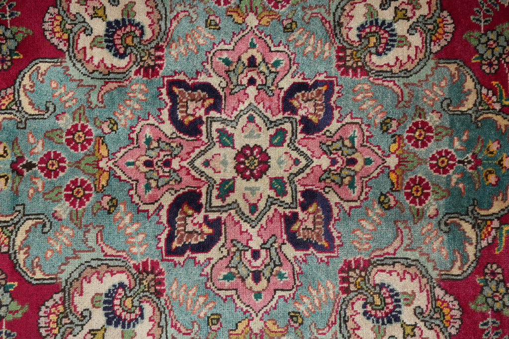 A close-up of a traditional patterned rug featuring a central floral medallion in pink, teal, cream, and burgundy with ornate vine motifs around it.