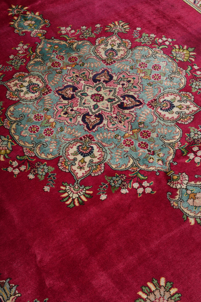 A close-up of a traditional Persian-style rug featuring a rich magenta red background with a large central medallion in light blue, pink, cream, and green floral motifs.