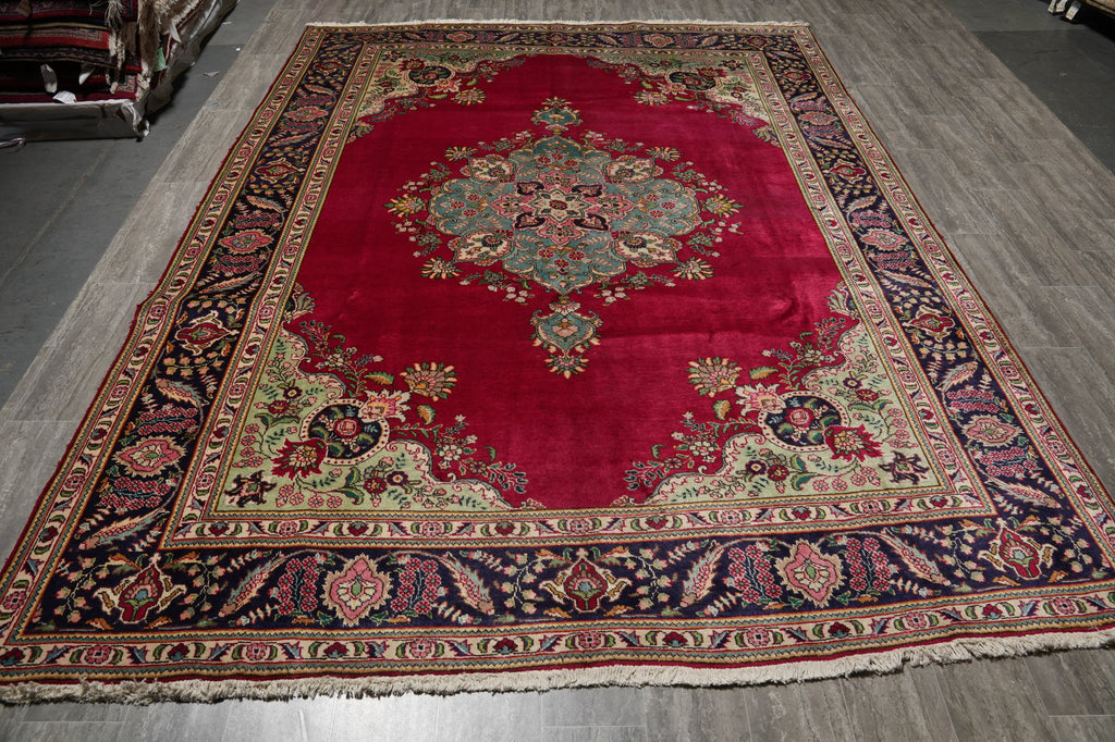 A large traditional Persian-style area rug with a deep red central field, ornate floral medallion, and detailed navy, beige, and gold borders, displayed on a gray wood floor.