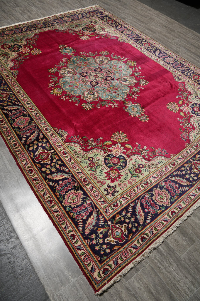A traditional oriental area rug with a vibrant red central field, an intricate multicolor floral medallion, and a dark navy blue border with beige and red details, laid out on a gray wood floor.