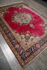 Load image into Gallery viewer, A traditional oriental area rug with a vibrant red central field, an intricate multicolor floral medallion, and a dark navy blue border with beige and red details, laid out on a gray wood floor.