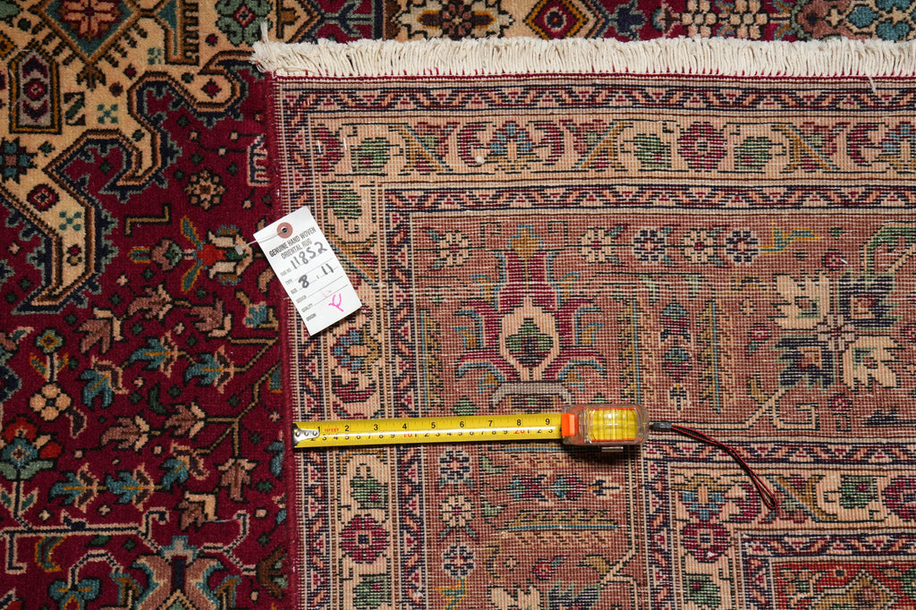 A detailed view of a traditional Persian-style area rug with a red and beige pattern, showing a white tag and a yellow tape measure laid out on the floor for scale.