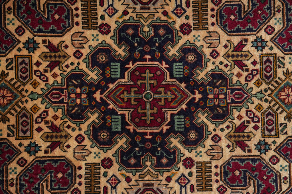A close-up of a traditional geometric patterned rug with a central medallion. The design features a warm beige background with prominent navy blue, burgundy red, and muted green motifs.