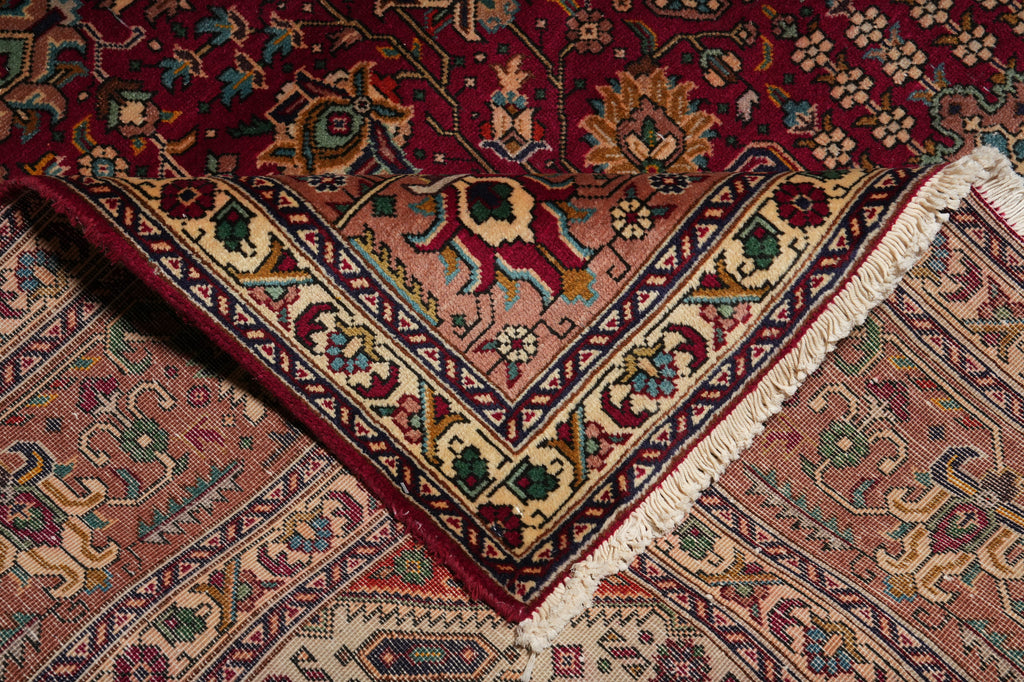 A close-up of a traditional patterned rug corner showing a deep red field, a beige and burgundy geometric border, cream fringe, and additional matching rug sections underneath.