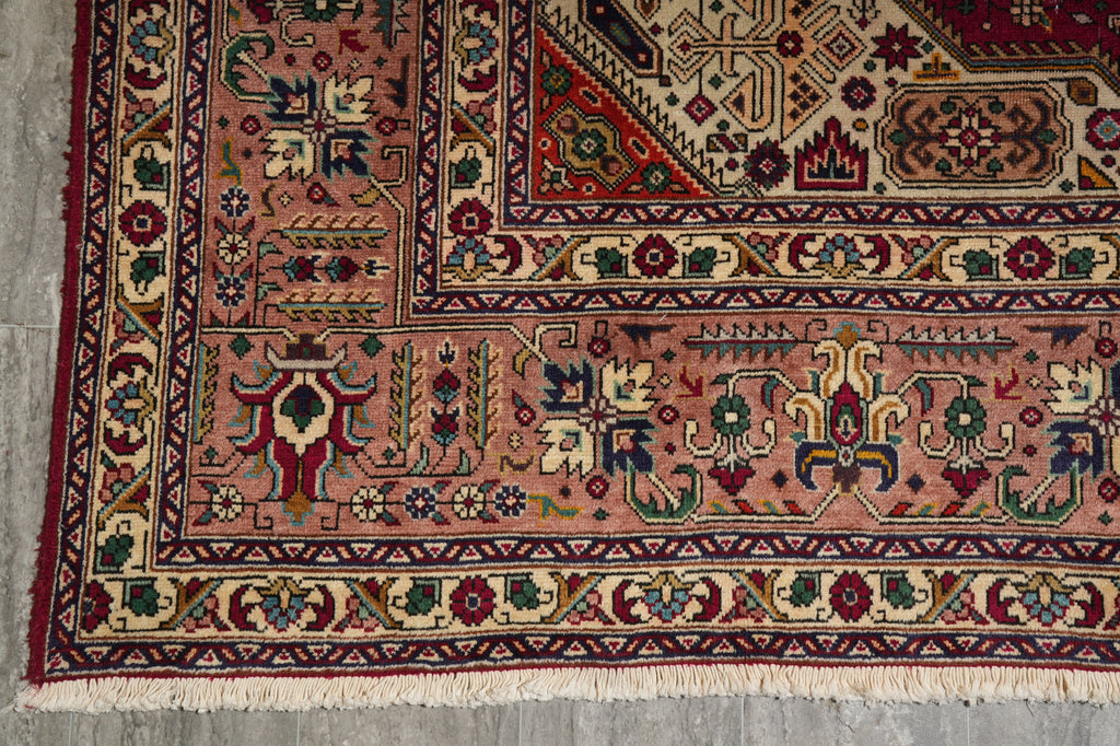 A close-up of a traditional patterned rug corner, showing a dusty pink field with red, navy, green, and beige geometric and floral motifs, finished with cream fringe.