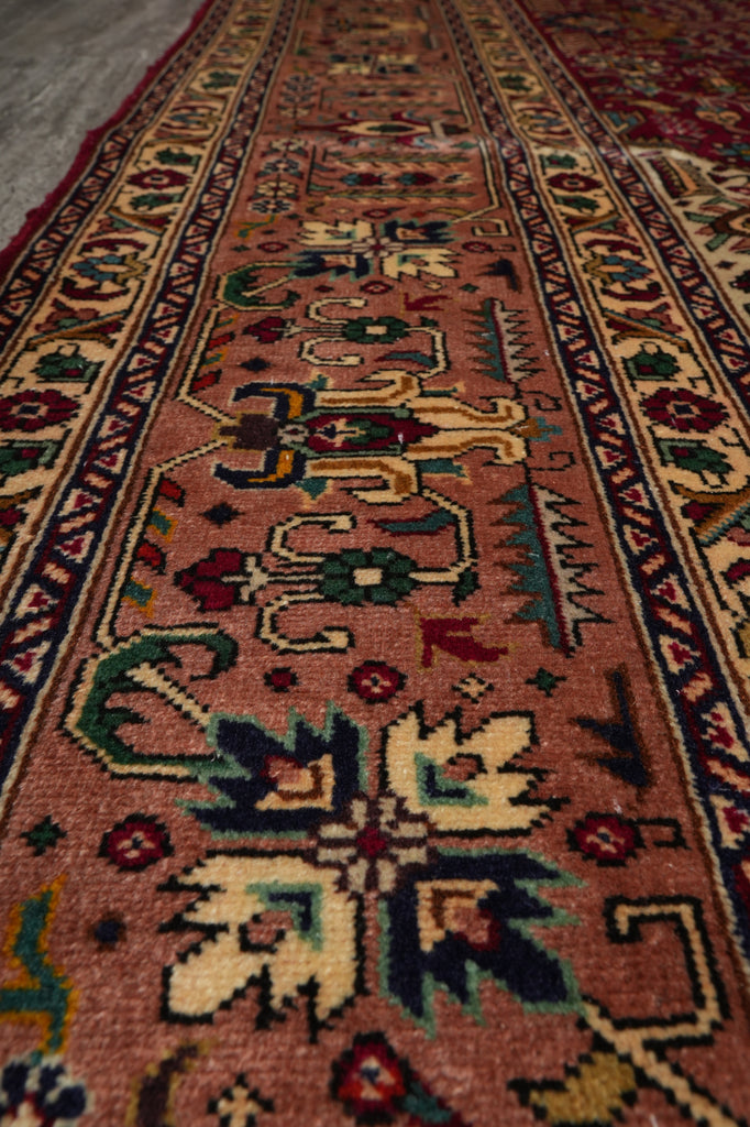 A close-up view of a traditional oriental runner rug with a dusty rose pink field, featuring intricate geometric and floral motifs in navy blue, green, beige, and red.