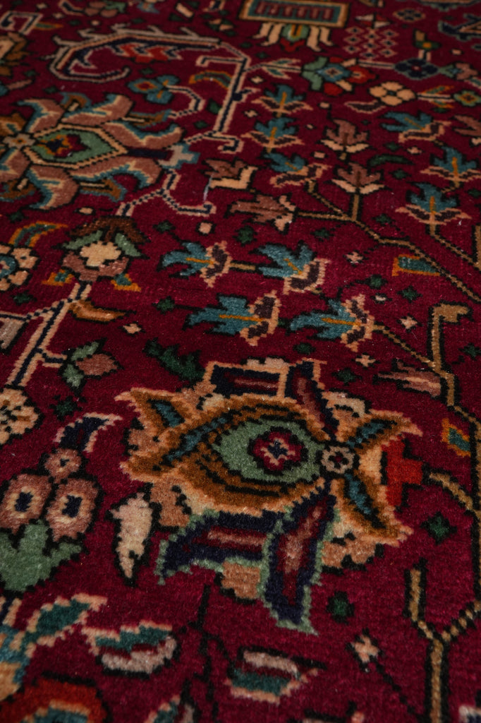 A close-up view of a traditional patterned rug with a deep red background and intricate floral motifs in beige, blue, green, and brown.
