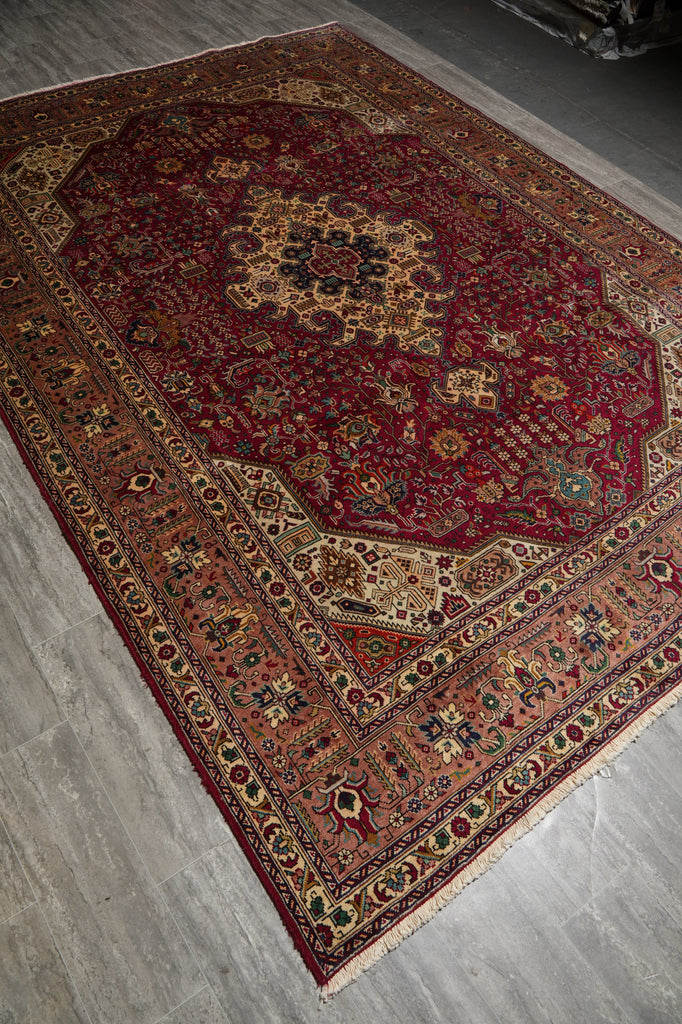 A traditional oriental area rug with a deep red central field, intricate floral motifs, and a detailed multi-colored border, shown laid out on a gray wood floor.