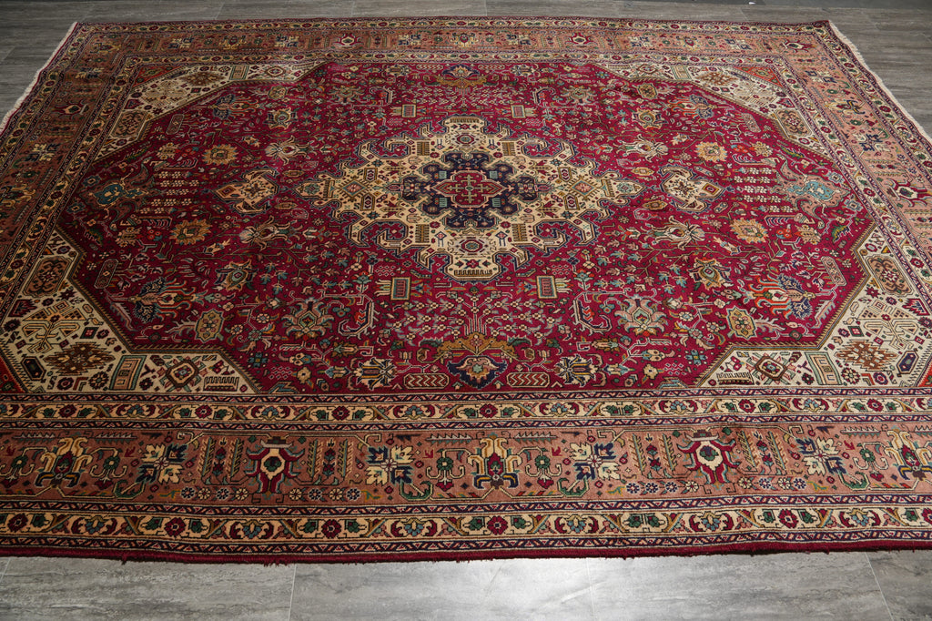 A large traditional Persian-style area rug with a deep red central field, intricate floral motifs, and detailed borders in beige, brown, blue, and green, laid out on a gray tiled floor.