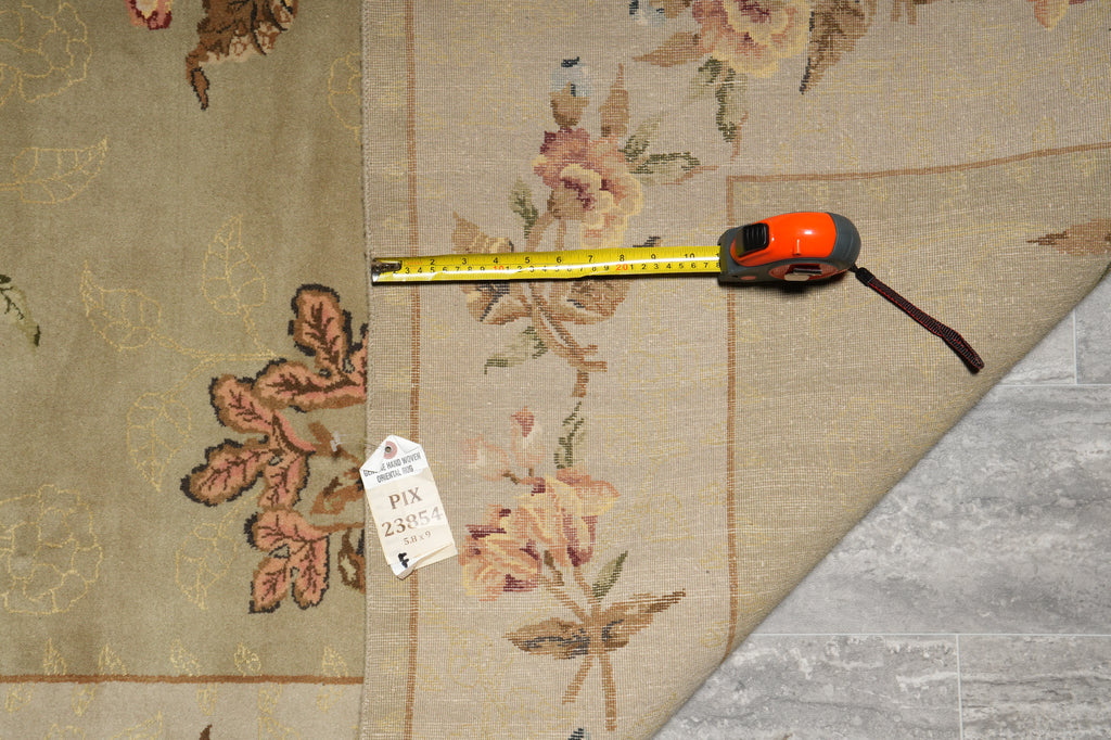 A close-up of a beige floral area rug with a tape measure and price tag placed on top, showing the fabric pattern and a folded corner revealing a gray floor underneath.