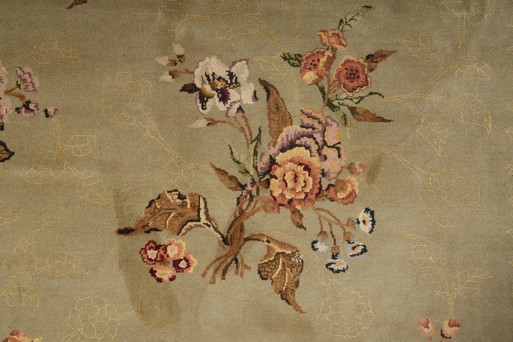 Close-up of a floral textile or rug pattern with embroidered-style flowers in peach, pink, cream, and muted green on a warm beige background with subtle gold-toned linework.